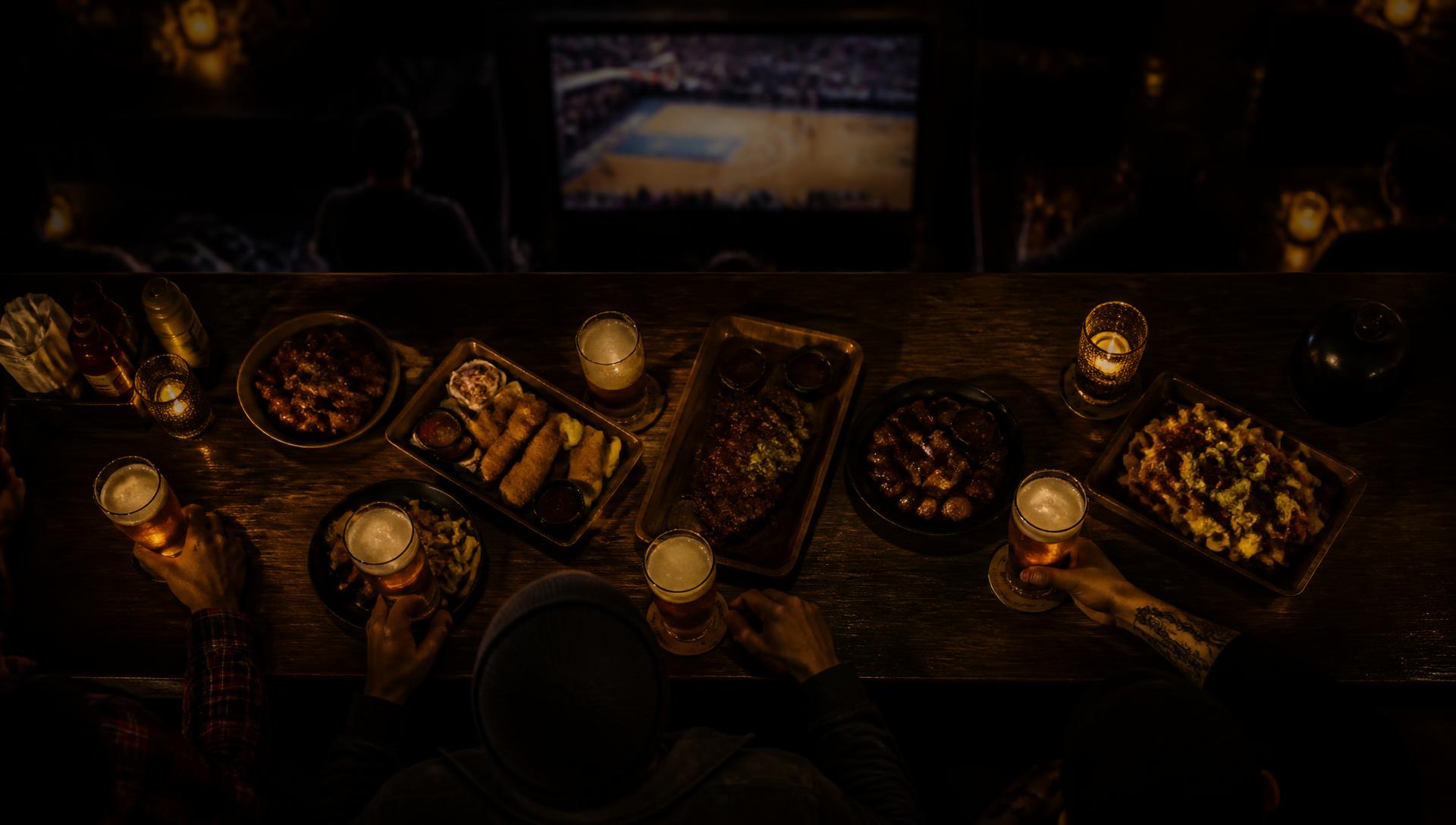 Mesa de bar vista de cima com copos de chopp, porções e um telão brilhando ao fundo